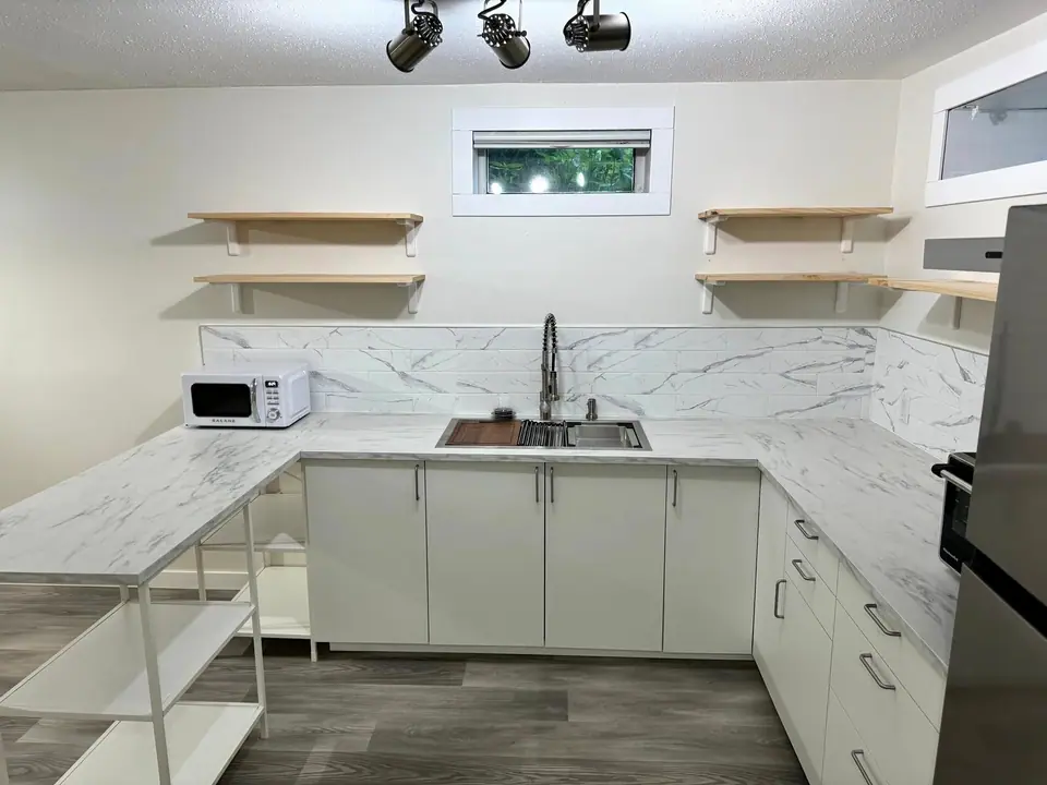 A finished white U-shaped kitchen with marble-look counters, open wood shelves and the sink under the window